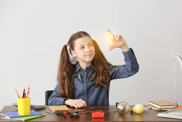 Cute little schoolgirl with glowing light bulb at table against grey background - Photo, Image