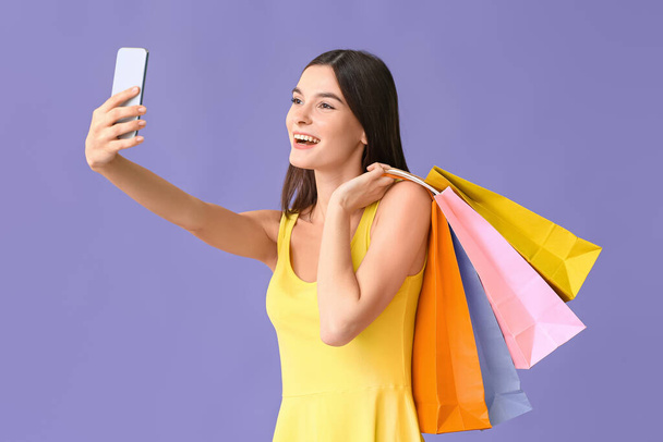 Mooie jonge vrouw met boodschappentassen nemen selfie op kleur achtergrond - Foto, afbeelding