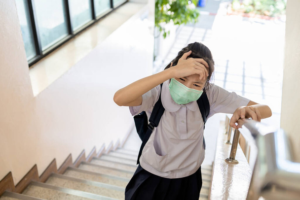 Fáradt ázsiai gyermek lány szenved fejfájás migrén, érinti fej vagy homlok kézzel, miközben sétál fel a lépcsőn az iskolában, tini diák egy maszk influenzát, fejfájás a stressz, betegség és láz - Fotó, kép