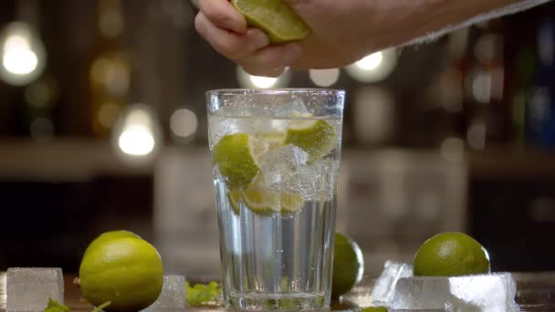 Close up male hand squeezing lime into a glass with tonic, soda water or cocktail. Glass with a refreshing drink, ice, limes and leaves on a table against the blurry lights background - Footage, Video