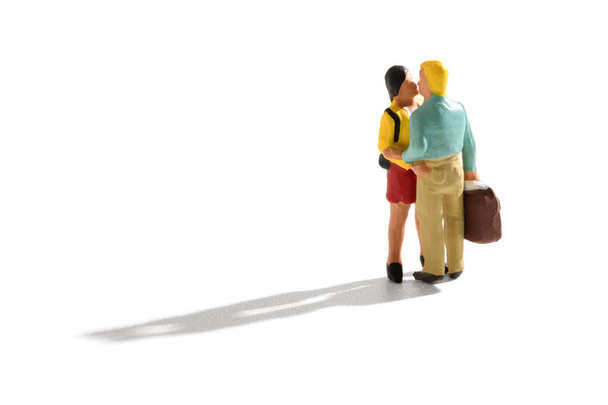 Miniatuur minnaars zoenen bij aankomst of vertrek met een jonge man en vrouw omhelzend als hij draagt een grote koffer voor reizen over wit met schaduw - Foto, afbeelding