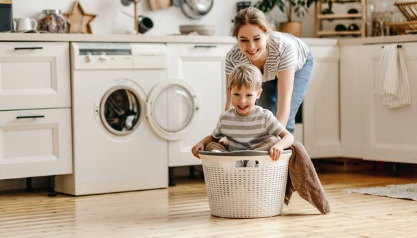 Cheerful young woman laughing and pushing basket with son while having fun in cozy kitchen in weeken - Photo, Image
