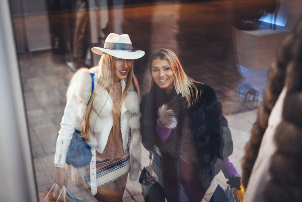 Zwei schöne, fröhliche Schwestern haben Spaß beim Einkaufen und zeigen durch das Schaufenster auf etwas. - Foto, Bild