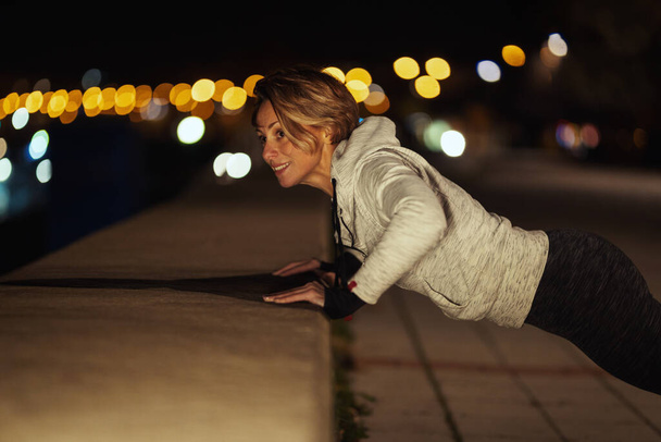 Young fitness woman is doing push-up exercise on the quay by the river in the evening. - Photo, Image