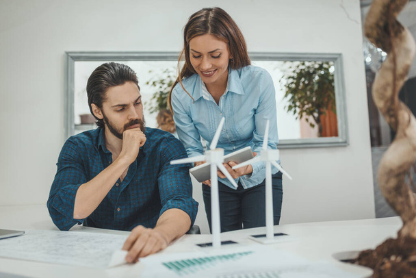 Умные прогрессивные молодые инженеры говорят о проекте ветряных мельниц в офисе, рядом со столом с симпатичными миниатюрными ветряными мельницами стоят на нем
. - Фото, изображение