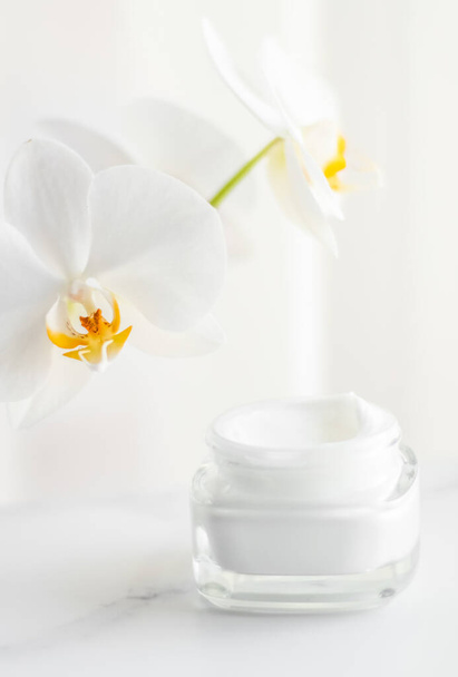 Увлажняющий крем для лица в банке и цветущий цветок орхидеи на белом фоне, косметика по уходу за кожей
 - Фото, изображение