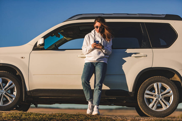 woman standing near white suv car surfing internet on the phone warm sunset light - Photo, image