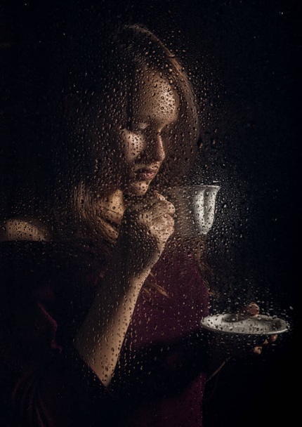 nice young girl drinks coffee behind the wet glass, raindrops on glass - Valokuva, kuva