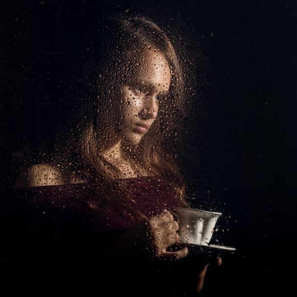 nice young girl drinks coffee behind the wet glass, raindrops on glass - Zdjęcie, obraz