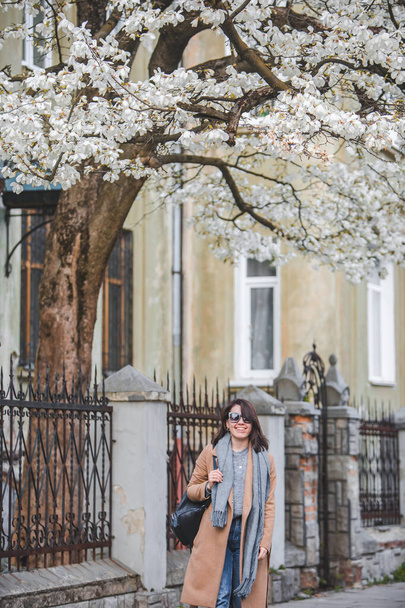 young stylish woman walking by city street in coat blooming tree on background. lifestyle - Foto, immagini
