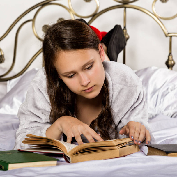 молодая студентка читает книгу, лежа на кровати, делая домашнее задание - Фото, изображение