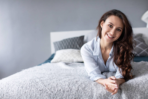 Portrait of beautiful young brunette woman with attractive smile in bed at home. Beautiful girl indoors. - Foto, afbeelding
