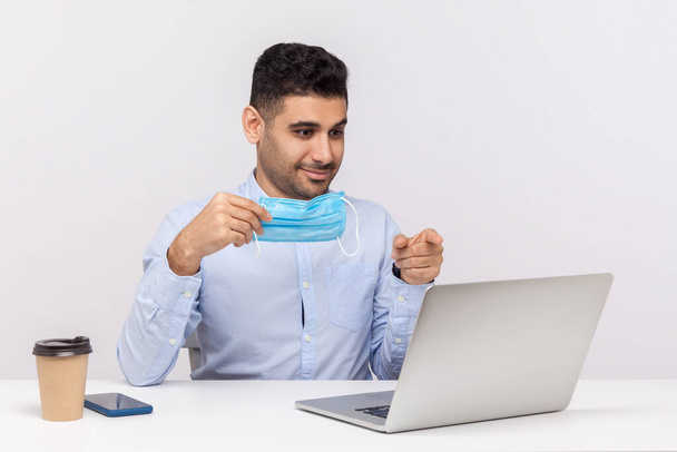 Positive office worker holding hygienic mask and pointing at laptop, watching medical program about contagious disease coronavirus, learning how to protect himself from 2019-nCoV. studio shot isolated - Foto, Imagen