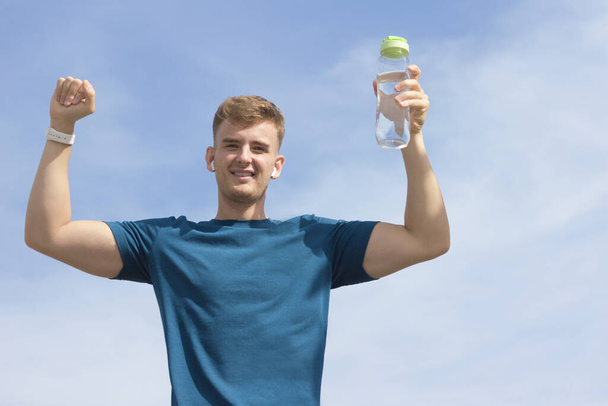 Happy handsome fit athletic guy with bottle of water, young man celebrating triumph, victory with his arms, hands raised up. Boy listening to music in wireless earphones, smiling. Successful athlete - Foto, immagini