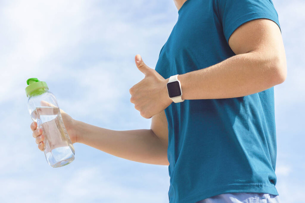 Close up photo of athlete's hand with smartwatch, bottle of water in hand. Unrecognizable person, fit man runner showing like gesture, thumb up. Healthy sport active fitness lifestyle, gadget concept - Foto, immagini