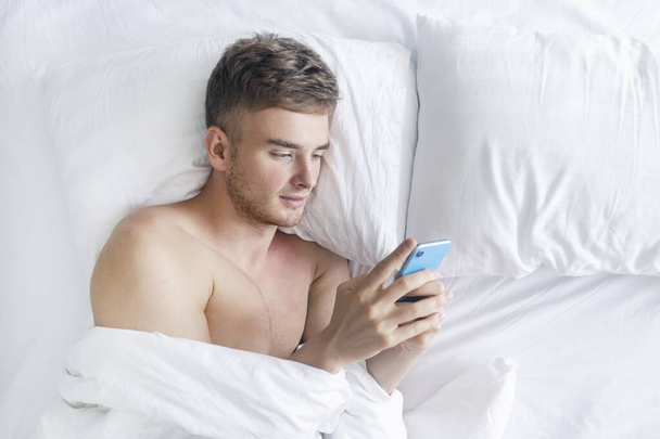 Handsome man, young guy lying in bed with cell mobile, using smart phone, typing, texting message or sms before sleep, fall asleep. Social media addiction. Love, relationships in the distance concept - Fotó, kép