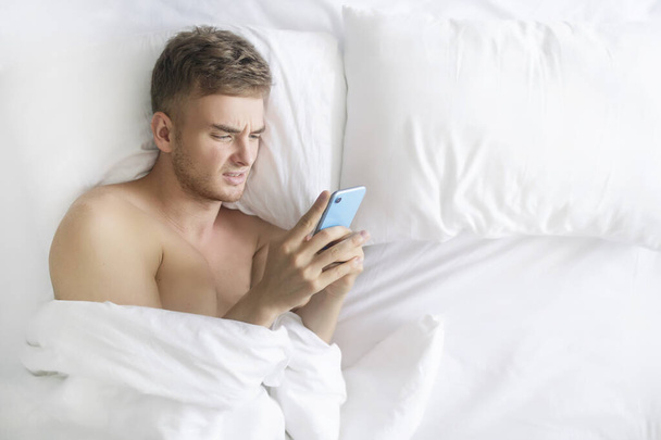 Handsome negative disgusted sad man, young upset angry tired guy lying in bed on pillow with cell mobile, using smart phone, get bad news, argue with wrinkled displeased face before sleep, sleeping - Fotó, kép