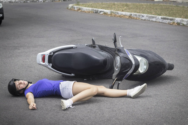 Downed biker in helmet from motorcycle, bike, moped. Girl, young unconscious woman lying on road, asphalt with broken leg injury. Accident with inattentive careless driver. Life threatening vehicle - Fotografie, Obrázek