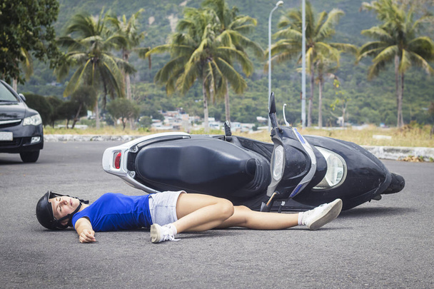 Downed girl with a motorcycle bike by a car lying unconscious on the road in helmet with broken leg. Accident on the road. Inattentive careless female biker crashed on vehicle. Dangerous situation. - Foto, imagen