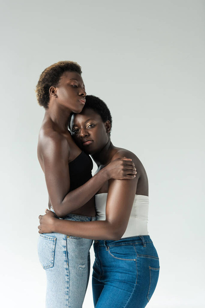 mooie Afrikaanse amerikaanse vrienden in jeans en tops knuffelen geïsoleerd op grijs  - Foto, afbeelding