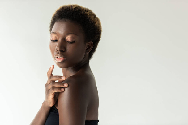 tender african american girl in black top touching shoulder isolated on grey - 写真・画像