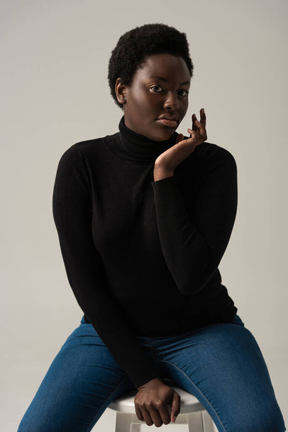 beautiful african american girl in jeans and black turtleneck sitting on stool isolated on grey - Fotoğraf, Görsel