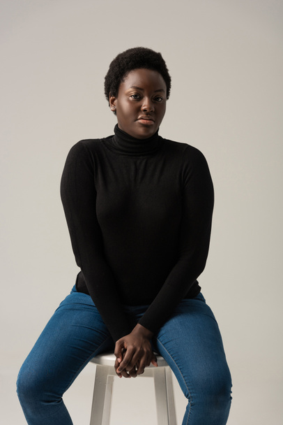 african american girl in jeans and black turtleneck sitting on stool isolated on grey - Foto, immagini