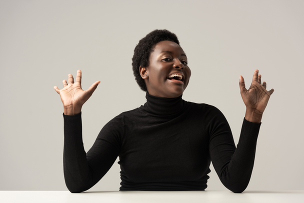 excited african american woman in black turtleneck sitting at table isolated on grey - Zdjęcie, obraz