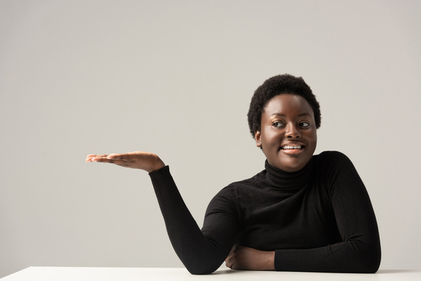 alegre afroamericana mujer en negro cuello alto sentado a la mesa y mostrando algo aislado en gris
 - Foto, imagen