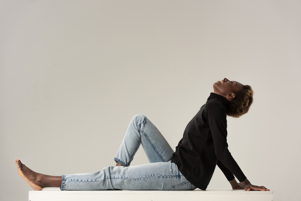 happy african american girl in jeans and black turtleneck sitting on white cube isolated on grey - Fotografie, Obrázek