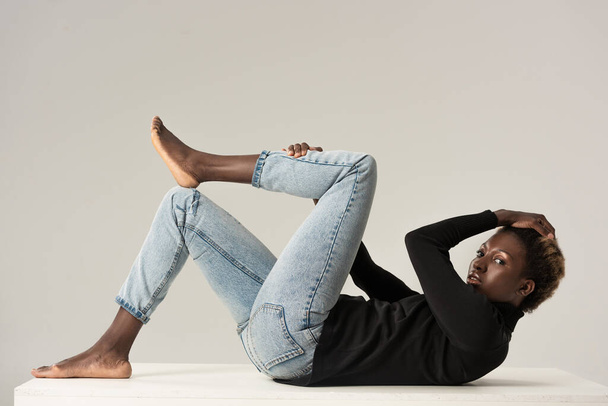 african american girl in jeans and black turtleneck sitting on white cube isolated on grey - Foto, afbeelding