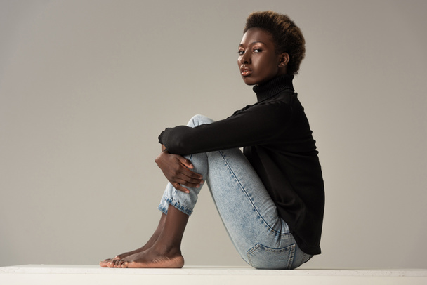 young african american woman in jeans and black turtleneck sitting on white cube isolated on grey - Foto, Imagem