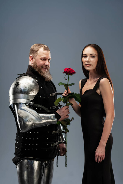 Όμορφο κορίτσι με μαύρο φόρεμα και ένα κόκκινο τριαντάφυλλο στο χέρι δίπλα σε έναν ιππότη με πανοπλία. - Φωτογραφία, εικόνα