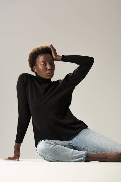 african american girl in black turtleneck sitting on white cube isolated on grey - Foto, afbeelding
