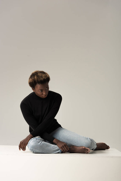 chica afroamericana en cuello alto negro sentado en cubo blanco aislado en gris
 - Foto, Imagen