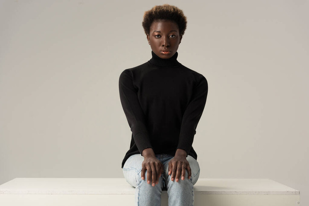 beautiful african american girl in jeans and black turtleneck sitting on white cube isolated on grey - Fotografie, Obrázek