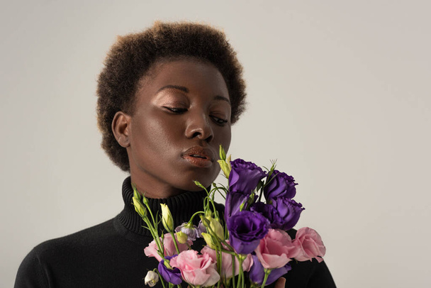 beautiful african american woman in black turtleneck holding eustoma flowers isolated on grey - Foto, afbeelding