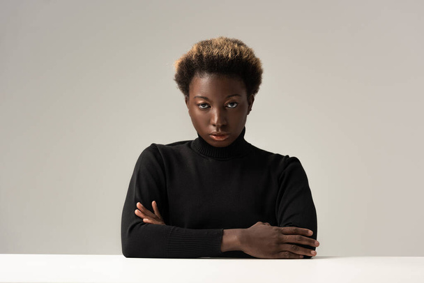 afro nainen musta poolokaula istuu pöydässä ristissä kädet eristetty harmaa
 - Valokuva, kuva