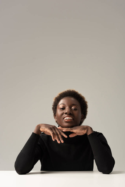 cheerful african american woman in black turtleneck sitting at table isolated on grey - Fotó, kép