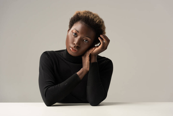 attractive tender african american girl in black turtleneck sitting at table isolated on grey - Фото, зображення