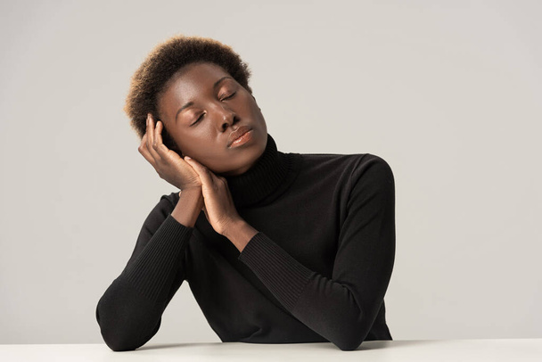 attractive tender african american woman with closed eyes in black turtleneck sitting at table isolated on grey - Photo, Image