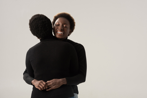 cheerful african american girlfriends in black turtlenecks hugging isolated on grey - Foto, Imagem