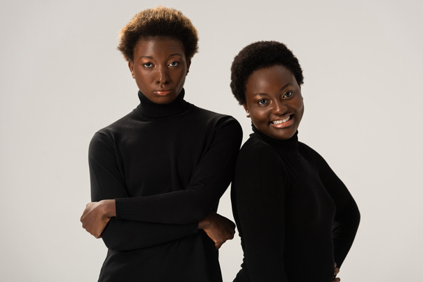 χαμογελαστές Αφροαμερικανίδες φίλες με μαύρα ζιβάγκο με σταυρωμένα χέρια, απομονωμένες σε γκρι. - Φωτογραφία, εικόνα