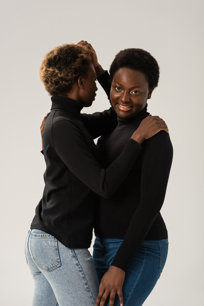 smiling african american women in black turtlenecks hugging isolated on grey - Photo, Image