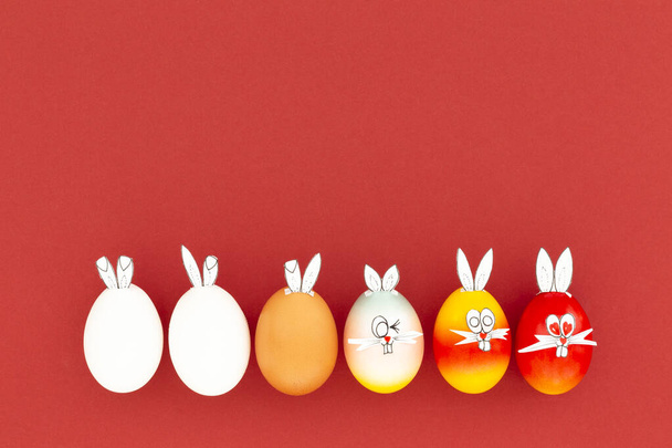 Uova di Pasqua dipinte bianche, marroni e colorate con divertenti orecchie da coniglio e volti dei cartoni animati su sfondo rosso
 - Foto, immagini