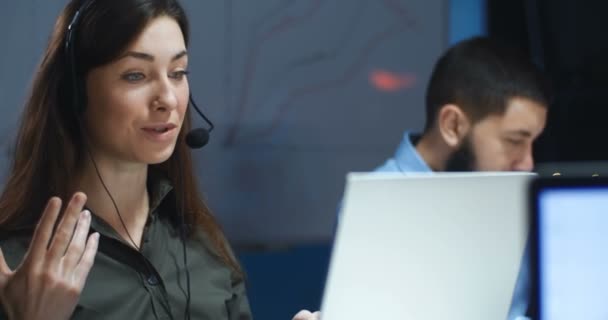Caucasian beautiful brunette young woman in headphones and mic having videochat at laptop computer in call center. Female worker of call-center speaking with client in evening. Male co-worker behind. - Кадры, видео
