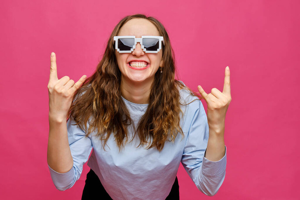 Κομψό καυκάσιος κορίτσι σε ένα απαλό μπλε t-shirt και 8-bit γυαλιά δείχνει ροκ χέρια σε ένα ροζ φόντο. Κλείσε.. - Φωτογραφία, εικόνα