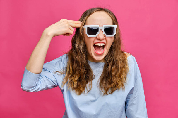 Stylish caucasian girl in a pale blue t-shirt and 8-bit glasses and an abstract gun from his hand pushed to her head smiling on a pink background. Close up. - Fotografie, Obrázek