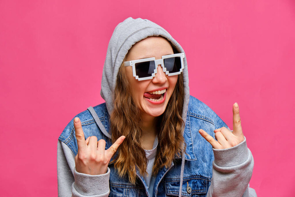 Elegante ragazza caucasica in jeans blu e occhiali a 8 bit e fa un segno rock con le mani che mostrano la lingua guardando la fotocamera su uno sfondo rosa. Da vicino.
. - Foto, immagini