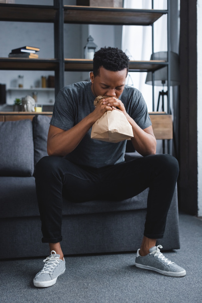 Worried african american man breathing with paper bag while having panic attack at home - 写真・画像